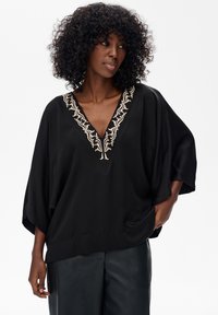 Schwarze Seidenbluse mit V-Ausschnitt, verziert mit aufwendiger beiger Stickerei entlang des Ausschnitts und weiten, fließenden Ärmeln.