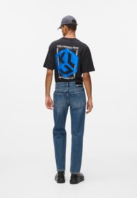 Juodas marškinėlis su mėlynu grafiniu spaudiniu, mėlyni džinsai ir juodi batai. Išskirtinis tekstas ant marškinėlių - "Karl Lagerfeld Jeans."