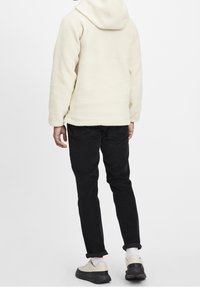 Crèmekleurige fleece hoodie met capuchon, gedragen met zwarte broek en lichtgekleurde schoenen. Zachte textuur en ontspannen pasvorm.