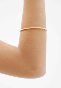 Una pulsera de bangle color dorado se encuentra sobre un brazo marrón claro, presentando un diseño suave y elegante con una forma redonda y delgada, además de sutiles acentos texturizados.