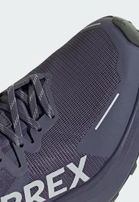 Zapatilla deportiva de color púrpura oscuro con parte superior de malla transpirable, puntera reforzada y letras de la marca en blanco. Presenta detalles reflectantes y suela texturizada.