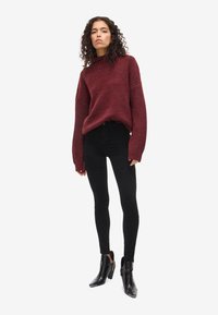 Burgundy kötött pulóver bordázott textúrával, túlméretezett ujjak, fekete, skinny farmerral és bokacsizmával párosítva. Minimalista dizájn, minta nélkül.