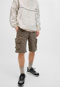 Beige windbreaker met zwarte piping, gecombineerd met olijfgroene cargoshorts met twee zijzakken, en zwarte sportschoenen met grijze accenten.