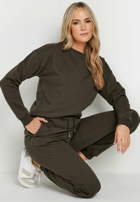 Sudadera verde oliva y joggers a juego hechos de tejido suave; cuenta con puños, cintura con cordón y bolsillos laterales. Combinado con zapatillas blancas.