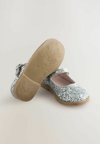 Scarpe glitterate argento con punta rotonda e dettaglio a fiocco. La suola testurizzata presenta un motivo floreale e di cervo. Fondo in gomma per maggiore aderenza.