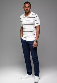 Vitrandig polotröja, mörkblå jeans och vita sneakers. Modellen står konfident med händerna i fickorna, kortärmad och med en avslappnad design.