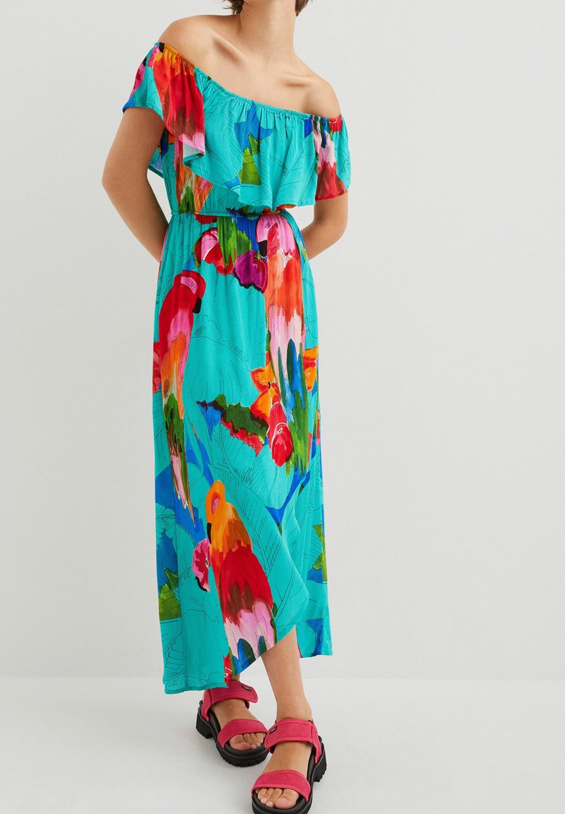 Robe maxi off-shoulder de couleur turquoise vif avec des impressions florales vibrantes. Tissu léger, décolleté froncé, associée à des sandales roses.
