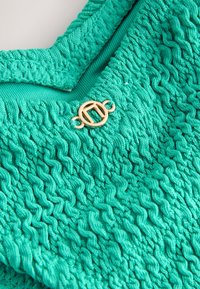 Gros plan sur un tissu tricoté turquoise texturé avec des motifs ondulés et un petit fermoir circulaire en or.