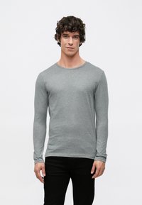 Langärmliges graues T-Shirt aus weichem Stoff, mit Rundhalsausschnitt und figurbetontem Schnitt. Kombiniert mit schwarzen Hosen. Minimalistischer Stil.