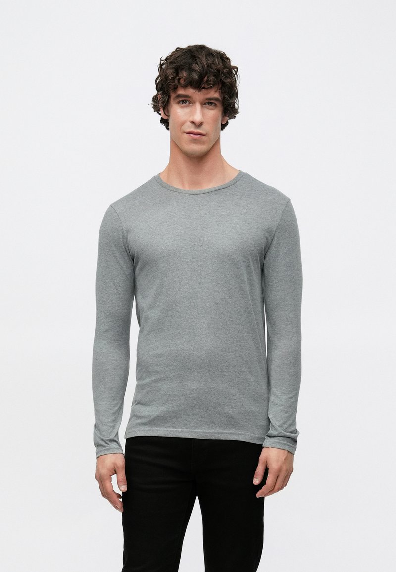 Langärmliges graues T-Shirt aus weichem Stoff, mit Rundhalsausschnitt und figurbetontem Schnitt. Kombiniert mit schwarzen Hosen. Minimalistischer Stil.