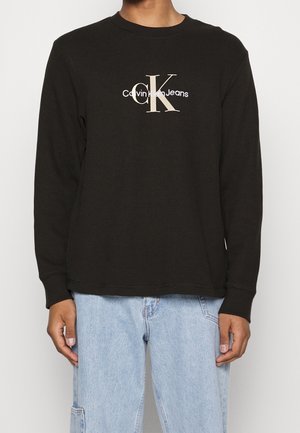 Person iført en sort langærmet Calvin Klein Jeans top med stort hvidt "cK" logo, kombineret med lyseblå denimjeans.