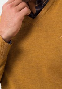 Pull de couleur moutarde, texture tricotée, col en V, poignets côtelés, avec un détail subtil de broderie, porté sur une chemise à carreaux.