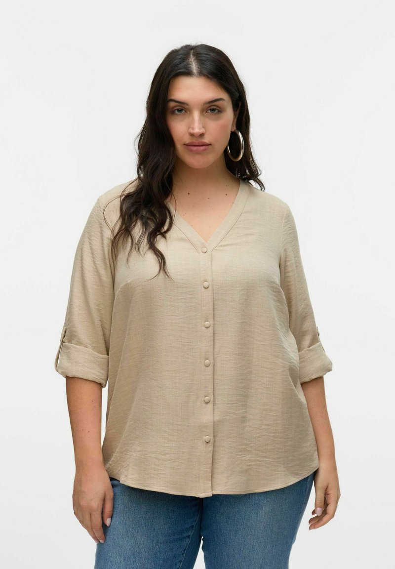 Beige overhemd met knopen en een V-hals, lange mouwen opgerold tot de ellebogen, en een gemêleerde stof. Gecombineerd met blauwe jeans.