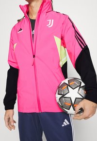 Lyserød og sort sportsjakke med lynlås foran, der har reflekterende gule detaljer og sorte ærmer. En fodbold med stjernemønster holdes.