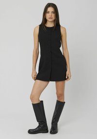 Robe noire sans manches avec un devant boutonné, coupe ajustée et deux poches latérales. Portée avec des bottes en cuir noires montantes.