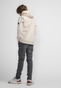 Felpa con cappuccio color beige chiaro, dotata di tasca frontale, abbinata a jeans slim-fit grigi e sneakers bianche con dettagli rossi.