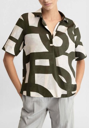 Donna che indossa una camicia a maniche corte con colletto con grandi motivi geometrici astratti in bianco e nero, abbinata a pantaloni grigio chiaro a righe sottili.