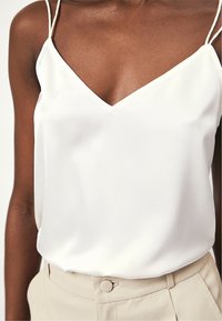 Witte satijnen camisole met een V-hals en dunne bandjes. Gladde textuur en een ontspannen pasvorm, gecombineerd met lichtbeige broeken met een knoopdetail.