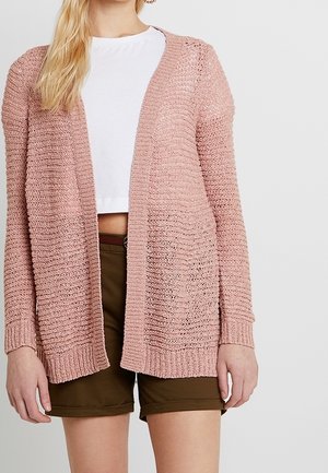Femme portant un cardigan ajouré rose clair, un t-shirt court blanc et un short vert olive, debout devant un fond gris uni.
