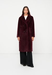 Burgundy double-breasted coat in soft, plush fabric with a wide collar, side pockets, and black buttons, paired with black wide-leg trousers.