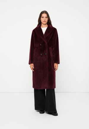 Manteau double boutonnage bordeaux en tissu doux et pelucheux avec un large col, des poches latérales et des boutons noirs, associé à un pantalon large noir.