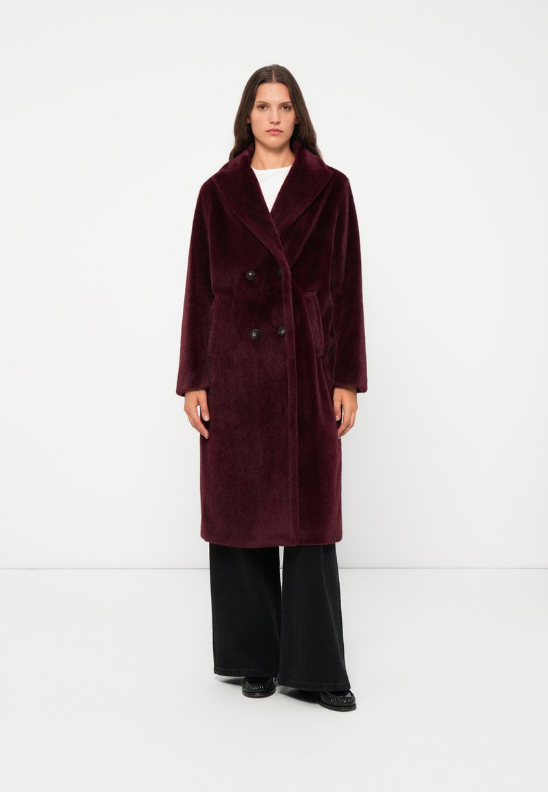 Burgundy double-breasted coat in soft, plush fabric with a wide collar, side pockets, and black buttons, paired with black wide-leg trousers.