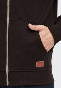 Bruine zip-up hoodie met een gestructureerde stof, zijzakken en een lederen label met het ingedeukte woord "BLEND".