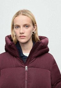 Chaqueta de plumas color marrón con un alto cuello y una capucha grande. Presenta una cremallera central y cierre de doble botón en el cuello. Textura suave.