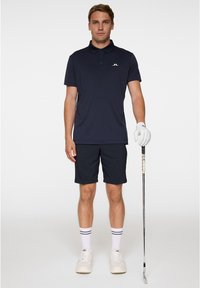 Marinblå polo med kort krage, ihop med mörka shorts. Outfitten kompletteras med vita strumpor med svarta ränder och vita sneakers.