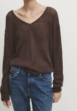 Persoon draagt een losse, donkerbruine gebreide V-hals trui, ingestopt in een high-waisted blauwe spijkerbroek met zichtbare voorzakken en lussen voor een riem.