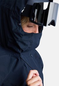 Czarna wodoodporna kurtka z wysokim kołnierzem, miękką teksturą i sznurkami, w zestawie z dużymi lustrzanymi goglami narciarskimi.