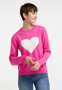 Pulover tricotat roz cu un design în formă de inimă albă, ribatată, în centru. Croială cu guler rotund și mansete ribatate, asortat cu blugi albaștri.