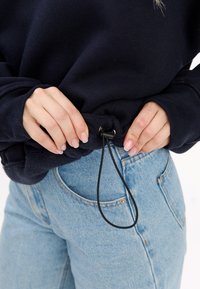 Granatowy sweatshirt z suwakiem na dole, w połączeniu z jasnoniebieskimi dżinsami. Zbliżenie na ręce regulujące suwakiem.