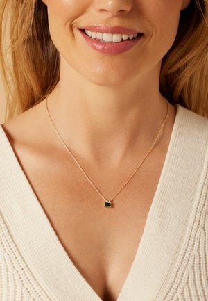 Gold chain necklace featuring a rectangular green stone pendant, worn with a cream v-neck sweater, highlighting the jewelry's simplicity.
