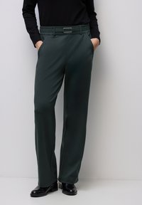 Mörkgröna, avsmalnande byxor med en slät textur, elastisk midja och en silverfärgad rektangulär spänne, kombinerade med svarta klackar.