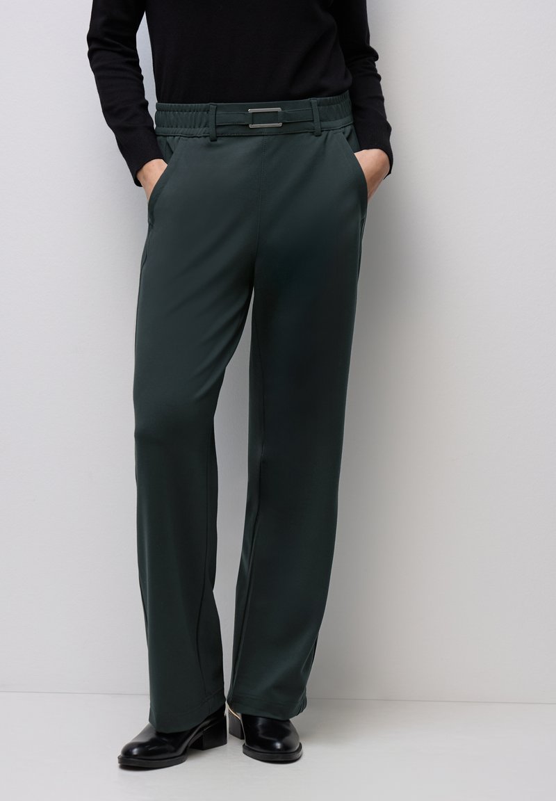 Mörkgröna, avsmalnande byxor med en slät textur, elastisk midja och en silverfärgad rektangulär spänne, kombinerade med svarta klackar.