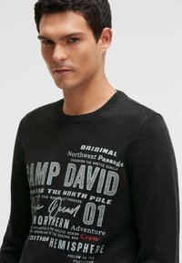 Schwarzer Strickpullover mit weißem und grauem Text: "CAMP DAVID", "Northern Adventure" und verschiedenen geografischen Referenzen auf der Vorderseite. Rundhalsausschnitt.