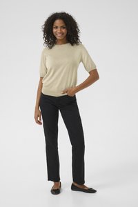 Nasmejana ženska s kodrastimi lasmi stoji z eno roko v žepu, oblečena v bež zgornji del s kratkimi rokavi, črne hlače in črni balerinki, na belem ozadju.