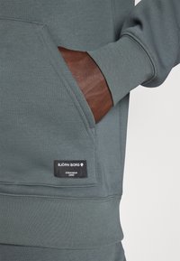 Sweatshirt verde-escuro com um bolso canguru e um detalhe em pele castanha. Apresenta uma etiqueta de tecido preta com a marca "BJÖRN BORG".