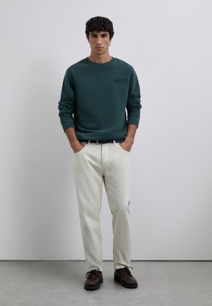 Joven vestido con un sudadera verde oscuro, pantalones blancos, cinturón negro y zapatos de cordones marrón oscuro, de pie contra un fondo gris liso.