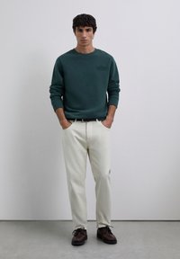 Jeune homme portant un sweat-shirt vert foncé, un pantalon écru, une ceinture noire et des chaussures marron foncé à lacets, se tenant devant un fond gris uni.