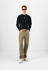 Sort langærmet skjorte med hvid syning og logo, parret med beige lige ben jeans og creme sneakers, modellen bærer sorte solbriller.