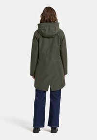 Olijfgroene waterdichte jas met capuchon, lange mouwen en een taps toelopende zoom. Gecombineerd met blauwe wijde jeans en zwarte schoenen.