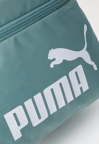 Mėlynas audinio maišelis, ant kurio yra balta Puma logotipas ir tekstas. Medžiaga turi tekstūruotą paviršių su kontrastingomis spalvomis ir užtrauktuko uždarymą.