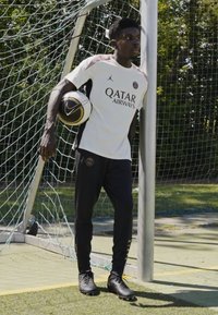 Vit t-shirts med korta ärmar och svarta detaljer samt texten "Qatar Airways", tillsammans med svarta träningsbyxor och svarta fotbollsskor, hållande en fotboll.