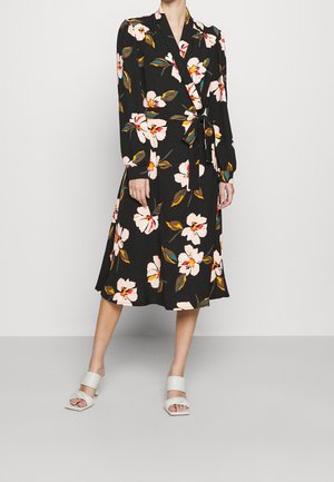 Woman wearing black midi wrap dress with large white and yellow floral print and white heeled sandals, standing against a plain background.