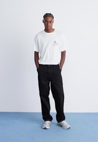 White cotton t-shirt with three circular graphic prints on the chest, paired with loose-fit black trousers and gray sneakers.