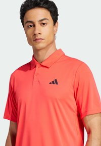 Polo de culoare corai strălucitor, cu guler, purtând un logo negru pe pieptul stâng, cu o textură netedă și o croială lejeră.