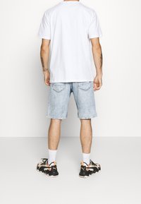 Camiseta blanca de manga corta, pantalones cortos de mezclilla azul claro y zapatillas deportivas negras con acentos naranjas. Tatuajes visibles en los brazos.