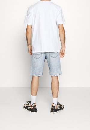 T-shirt blanc à manches courtes, short en denim bleu clair et chaussures de sport noires avec des accents orange. Tatouages visibles sur les bras.